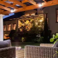 Drewniana pergola z lampami sufitowymi zewnętrznymi oświetla taras z meblami rattanowymi i ogrodem.