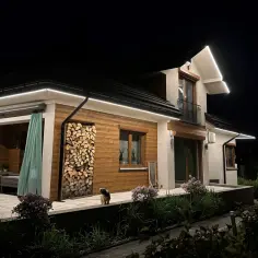 Elewacja domu oświetlona od dołu, podkreślająca drewniane elementy i stos drewna. Mały piesek stoi na tarasie.