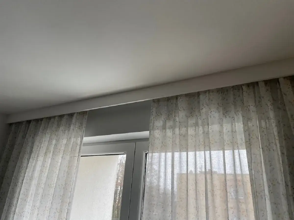 Jasne, oto propozycja:

Jasny, kwiecisty materiał zasłania okno, a nad nim widać gruby pasek pod sufitem.